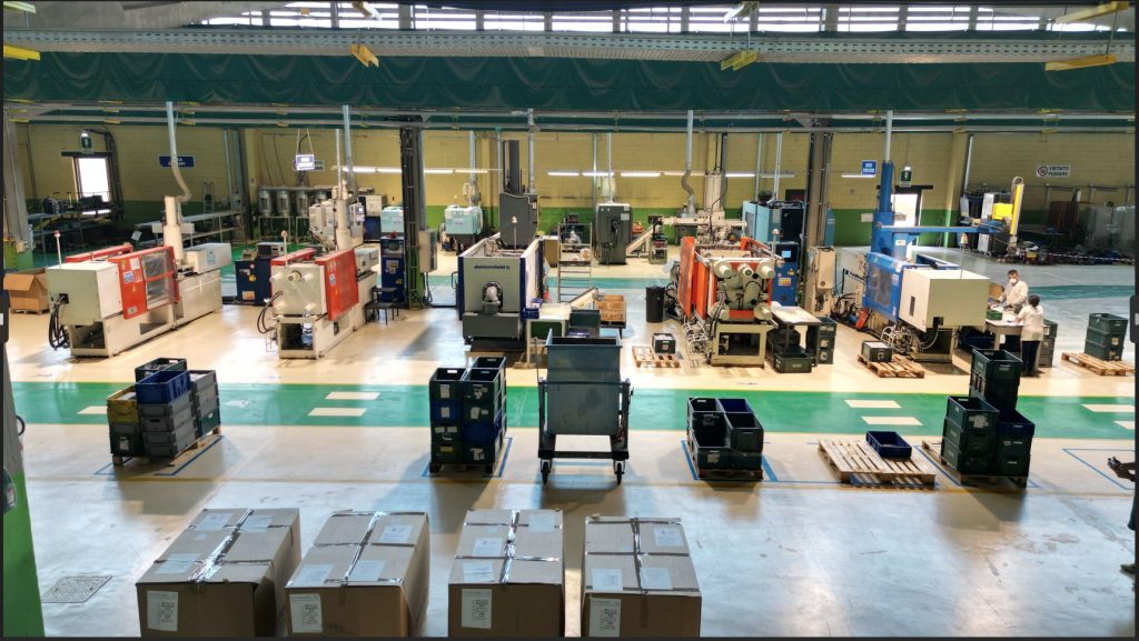 Area Produttiva e Macchine Interno di un reparto produttivo con diverse presse a iniezione bianche e arancioni allineate. Operatori lavorano sui macchinari lungo corridoi verdi delimitati.