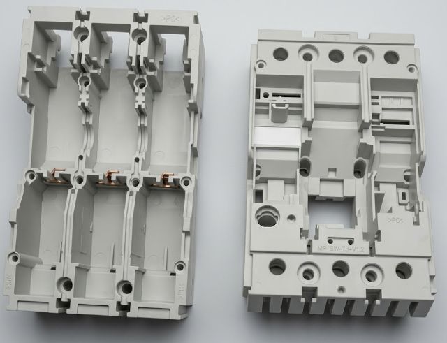Scocche per Dispositivi Elettrici Due grandi basi in plastica grigia per dispositivi elettrici affiancate. Quella a sinistra mostra profonde cavità con inserzioni in rame, quella a destra espone fori di montaggio e nervature strutturali.