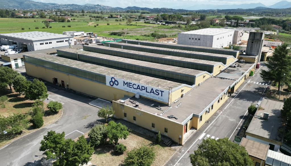 Il Quartier Generale MecaPlast Vista aerea del capannone industriale. Sulla lunga facciata gialla dell'edificio spicca una grande insegna con il logo MecaPlast e la scritta 'plastic components from Italy'